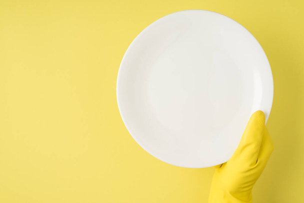 Top view photo of hand in yellow rubber glove holding white clean plate on isolated yellow background with blank space - 写真・画像