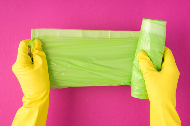 Πάνω όψη φωτογραφία των χεριών σε κίτρινα γάντια ξεδιπλώνοντας πράσινες σακούλες σκουπιδιών σε απομονωμένο ροζ φόντο με copyspace - Φωτογραφία, εικόνα