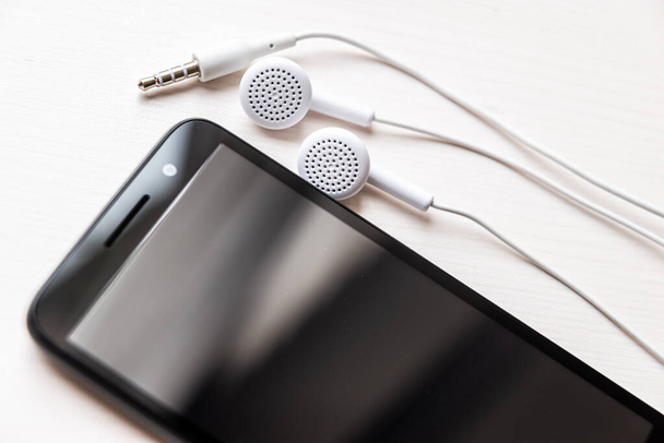Smartphone με συνδεδεμένα ακουστικά, λευκό χρώμα. Ακουστικό καλώδιο τυλιγμένο γύρω από το τηλέφωνο. Χρήση ακουστικών για να ακούτε μουσική ή για να κάνετε τηλεφωνήματα. - Φωτογραφία, εικόνα