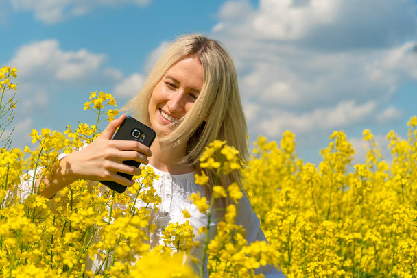 Young beautiful smiling blonde woman in a white shirt takes a selfie in a rapeseed field on a sunny day against a blue sky with clouds - Fotografie, Obrázek