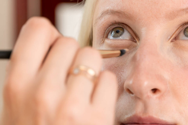Makijaż artysta używa pędzla do zastosowania ukrywacza pod oczami młodej kobiety. Proces makijażu - Zdjęcie, obraz