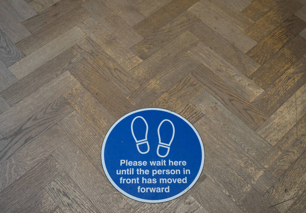 A round floor sign with white text on blue background and two footprint symbols.It says 'please wait here until the person in front has moved forward' .Coronavirus pandemic - Photo, Image
