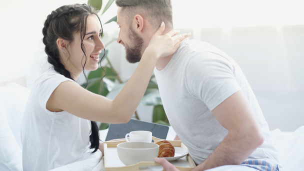 cheerful girlfriend with braids hugging happy man bringing breakfast tray in bed - Photo, Image