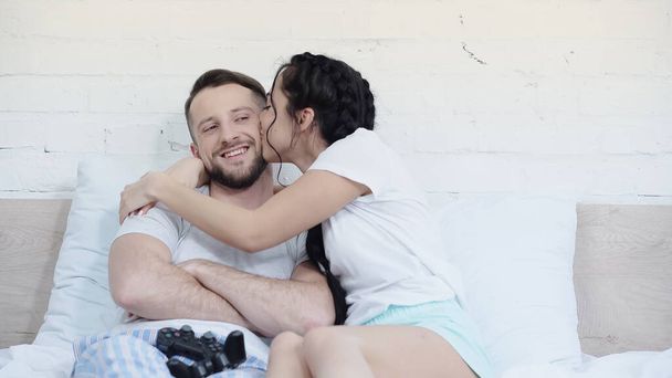 KYIV, UKRAINE - JUNE 17, 2021: young woman kissing cheerful man near joysticks on bed - Photo, Image