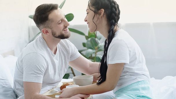 happy brunette woman with braids bringing breakfast tray to boyfriend in bed - Photo, Image