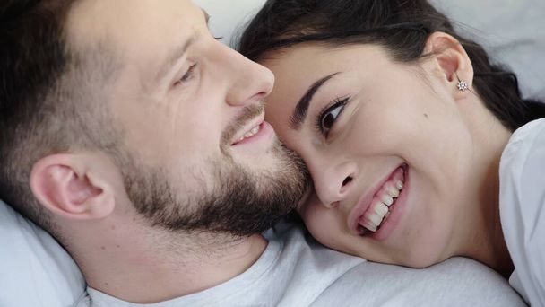 joyful brunette woman leaning on bearded boyfriend while resting on bed - Φωτογραφία, εικόνα