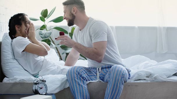 happy bearded man holding red box with wedding ring while doing proposal to girlfriend in bedroom  - Photo, image