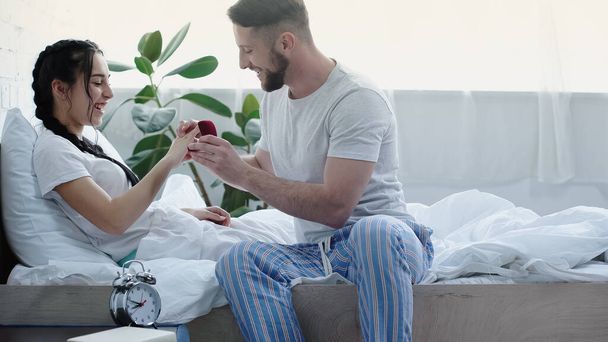 happy bearded man holding red box while wearing wedding ring on finger of girlfriend in bedroom  - Photo, image