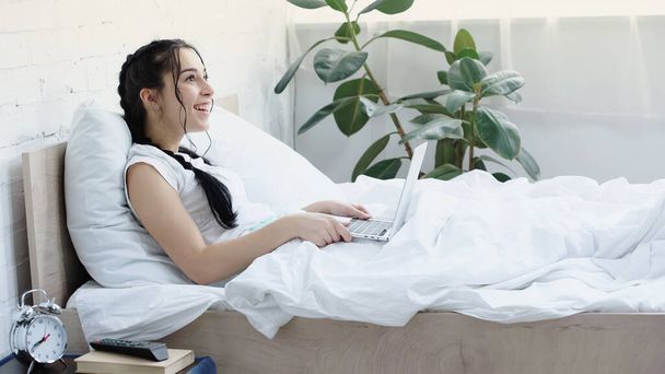 morena freelancer sonriendo mientras usa el portátil en la cama - Foto, imagen