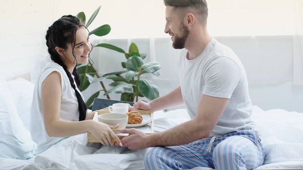 szczęśliwy człowiek przynosząc tacę śniadaniową do uśmiechniętej dziewczyny z warkoczami w sypialni  - Zdjęcie, obraz