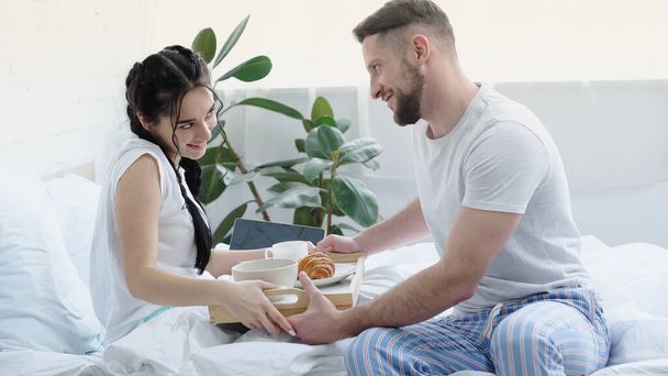 happy man bringing breakfast tray to joyful girlfriend with braids in bedroom  - Фото, зображення