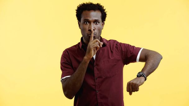 curly african american man in burgundy shirt and wristwatch showing hush sign isolated on yellow - Photo, Image