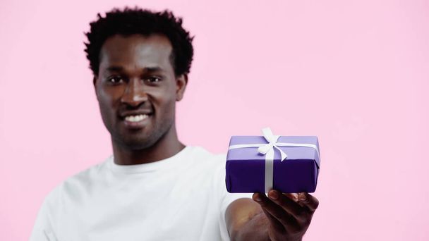 verschwommen und glücklich afrikanisch-amerikanischer Mann in weißem T-Shirt mit eingewickeltem Geschenk isoliert auf rosa - Foto, Bild