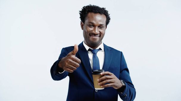 pleased african american businessman holding paper cup and showing thumb up isolated on blue - Fotó, kép