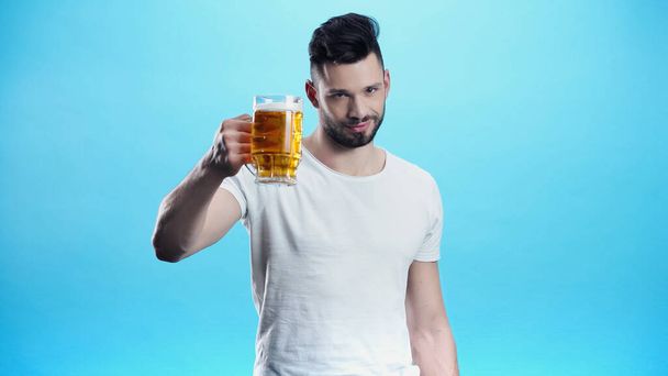complacido hombre sosteniendo taza de cerveza fría aislada en azul - Foto, imagen