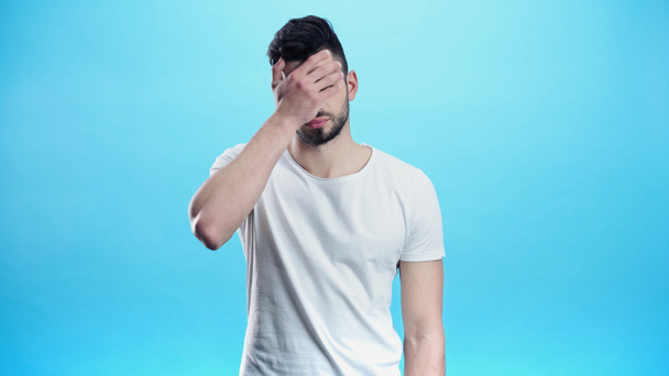 depressed man in white t-shirt covering eyes with hand isolated on blue - 写真・画像