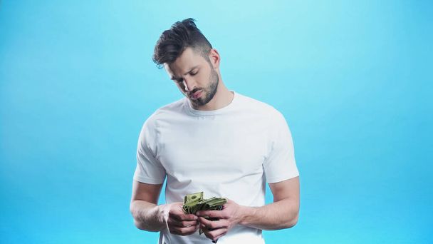 fiatal férfi fehér pólóban számolás dollár elszigetelt kék - Fotó, kép