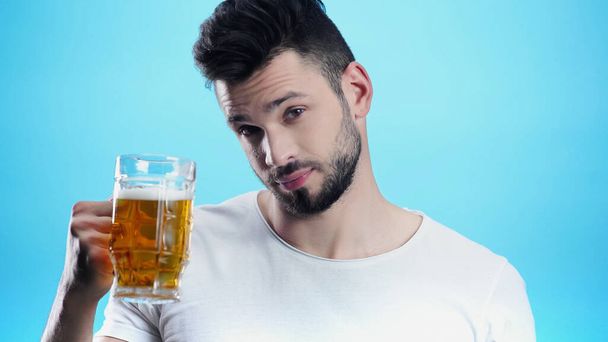 young man looking at camera while holding mug of beer isolated on blue - Fotoğraf, Görsel