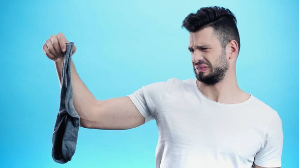 displeased man grimacing while holding smelly sock isolated on blue - Φωτογραφία, εικόνα
