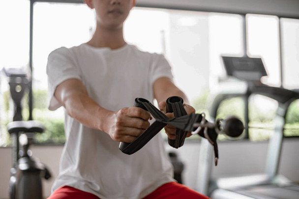 Concepto de gimnasio de entrenamiento un adolescente masculino usando un equipo de gimnasio tirando de sus dos brazos musculares contra la máquina. - Foto, imagen