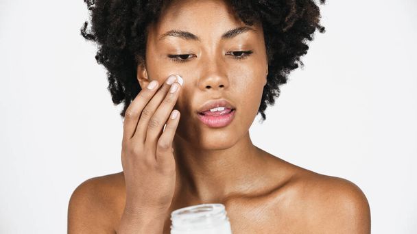 Mujer afroamericana con piel perfecta aplicando crema cosmética en la cara aislada en gris  - Foto, Imagen