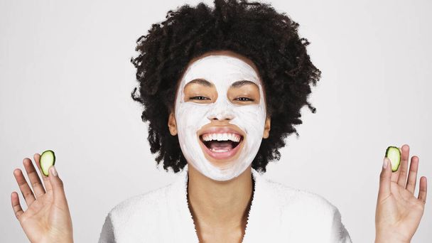 Fröhliche afrikanisch-amerikanische Frau mit Gesichtsmaske hält Gurkenscheiben isoliert auf grau  - Foto, Bild