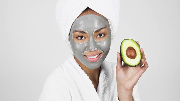 Mladá africká americká žena s hliněnou maskou na obličeji a ručníkem na hlavě drží avokádo izolované na šedé  - Fotografie, Obrázek
