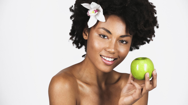 Uśmiechnięta Afroamerykanka z orchideą we włosach trzymająca zielone jabłko odizolowane na szaro  - Zdjęcie, obraz