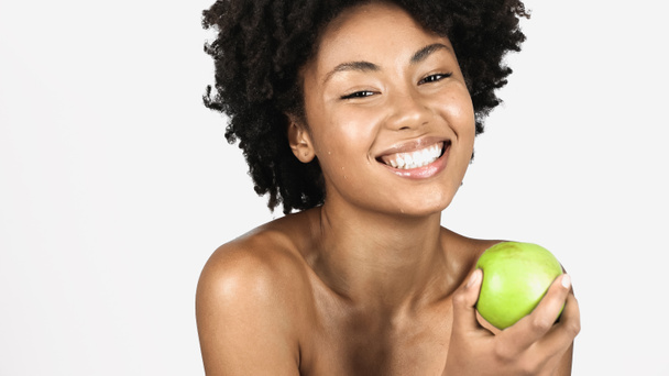Positive african american woman with naked shoulders holding juicy green apple isolated on grey  - 写真・画像