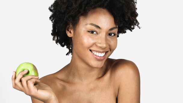 Uśmiechnięta Afroamerykanka z czystą skórą patrząca w obiektyw i trzymająca zielone jabłko odizolowane na szaro  - Zdjęcie, obraz