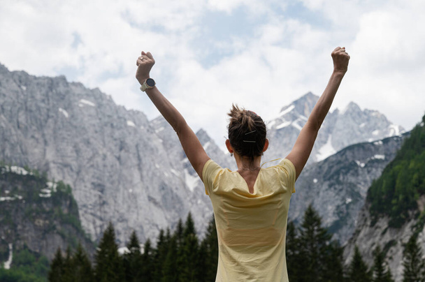 View from behind of a young woman standing under beautiful rocky mountains with her arms raised high. - Photo, Image