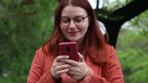 Usmívající se běloška v brýlích, držící smartphone za použití mobilní aplikační techniky venku. Šťastná mladá žena textové kontroly online sociálních sítí aplikací v městském parku. - Záběry, video