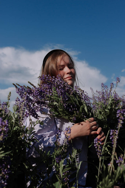 Mujer joven sonriente en camisa sosteniendo ramo de flores púrpuras de pie solo entre el campo frente al cielo azul disfrutando de la luz del sol a la hora dorada. Traje de verano. Chica con flores y ojos cerrados - Foto, Imagen