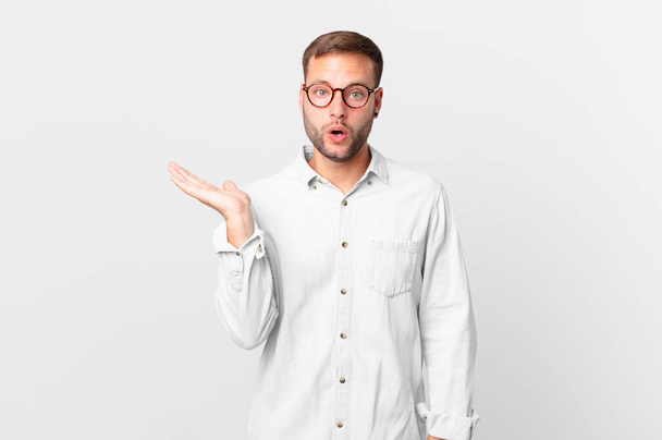 handsome blonde man looking surprised and shocked, with jaw dropped holding an object - 写真・画像