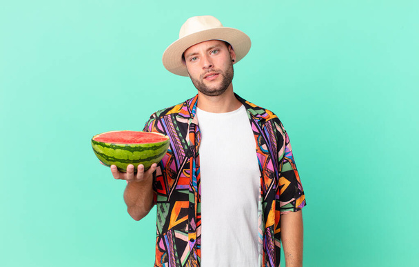 handsome traveler man feeling puzzled and confused and holding a watermelon. holidays concept - Foto, imagen