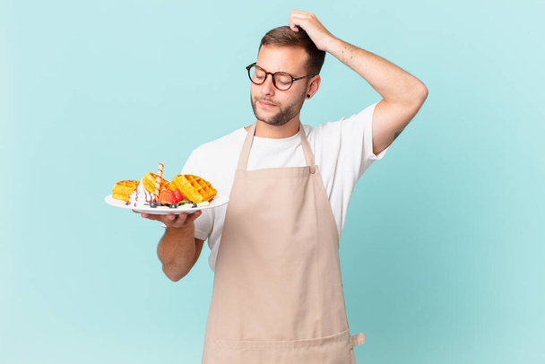 young handsome blonde man feeling puzzled and confused, scratching head. cooking waffles concept - Foto, Imagen
