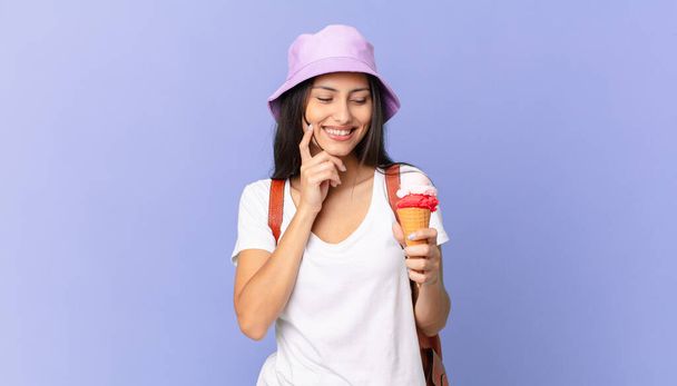pretty hispanic tourist smiling with a happy, confident expression with hand on chin and holding an ice cream - Foto, afbeelding