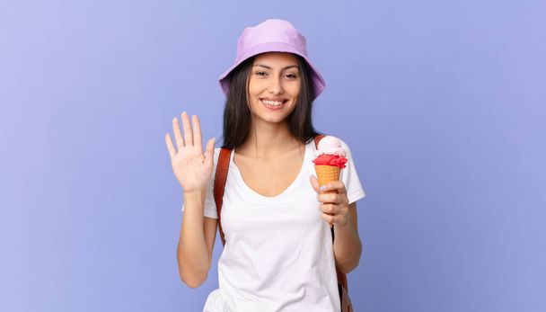 pretty hispanic tourist smiling happily, waving hand, welcoming and greeting you and holding an ice cream - Photo, image