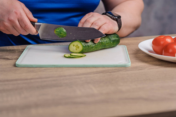 Un uomo taglia un cetriolo su una tavola. Pomodoro e cipolle verdi si trovano su un piatto bianco - Foto, immagini