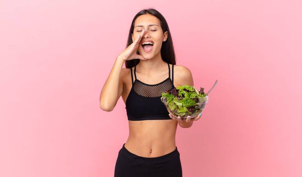 hispanische Fitness-Frau fühlt sich glücklich, gibt einen großen Schrei mit den Händen neben dem Mund und hält einen Salat - Foto, Bild