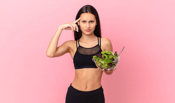 hispanic fitness woman feeling confused and puzzled, showing you are insane and holding a salad - Foto, Bild