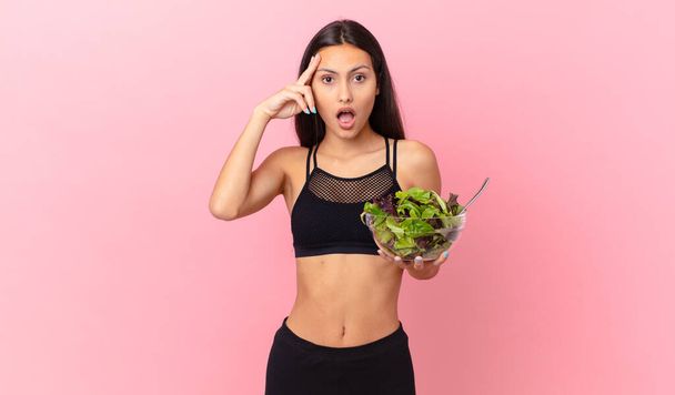 hispanic fitness woman looking surprised, realizing a new thought, idea or concept and holding a salad - Photo, Image