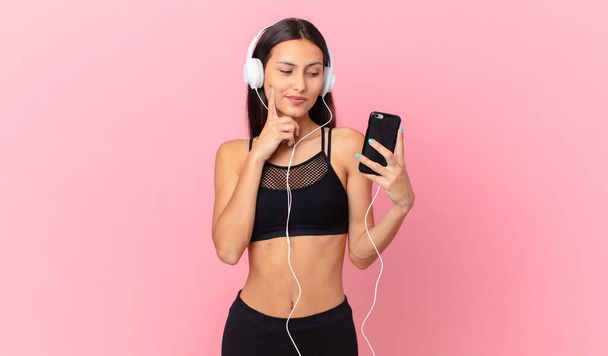 hispanic fitness woman smiling happily and daydreaming or doubting with headphones and a phone - Photo, image