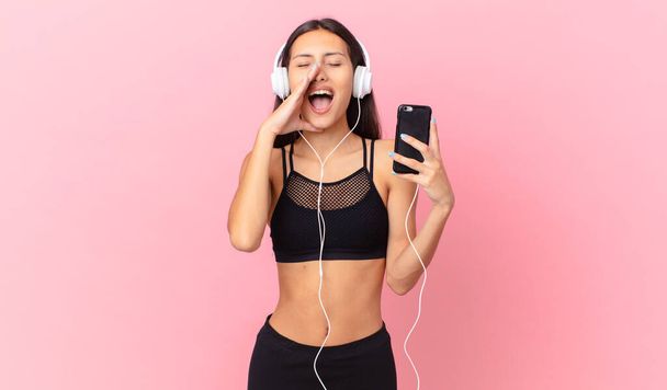 hispanic fitness woman feeling happy,giving a big shout out with hands next to mouth with headphones and a phone - Zdjęcie, obraz