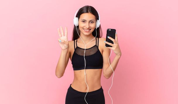 hispanic fitness woman smiling and looking friendly, showing number three with headphones and a phone - Foto, Imagen
