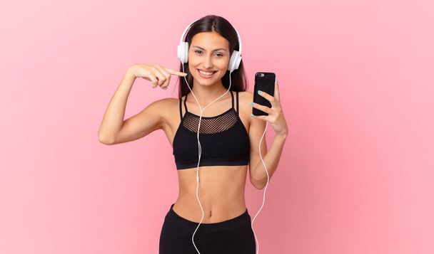 hispanic fitness woman smiling confidently pointing to own broad smile with headphones and a phone - Фото, изображение