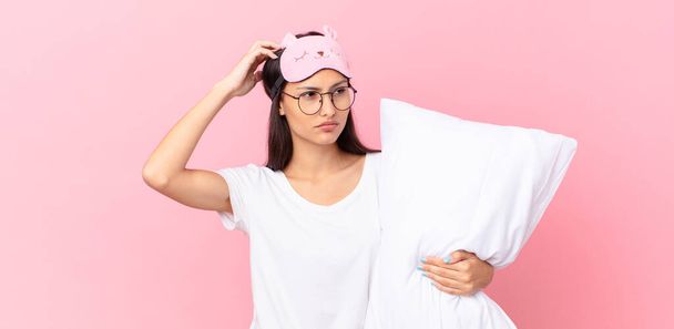 hispanic woman wearing pajamas feeling puzzled and confused, scratching head and holding a pillow - Photo, image
