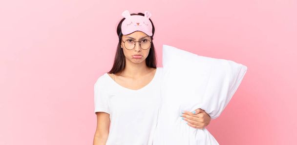 hispanic woman wearing pajamas feeling sad and whiney with an unhappy look and crying and holding a pillow - Фото, изображение
