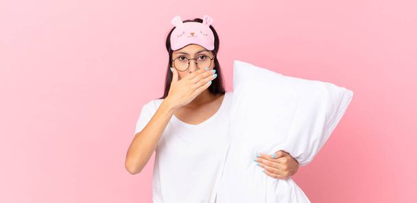 hispanic woman wearing pajamas covering mouth with hands with a shocked and holding a pillow - Foto, imagen