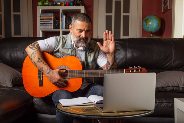 Uomo moderno seduto sul divano a casa saluta l'insegnante lezioni di chitarra online. Concetto di apprendimento online. - Foto, immagini
